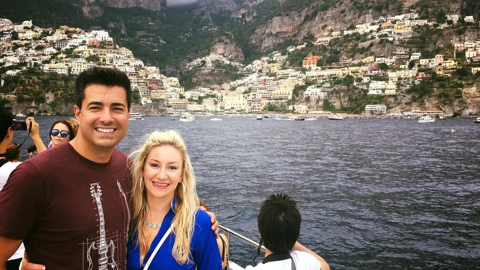 Alex e Kristin juntos num barco com a orla e a encosta de Positano atrás