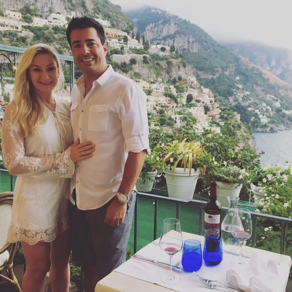 Alex e Kristin junto a uma mesa de terraço com taças e vista da encosta de Positano e do mar