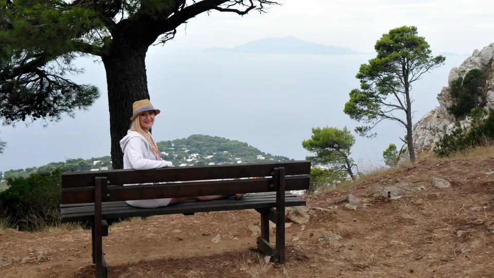 Kristin sitting on a wooden bench beneath a pine tree with the sea and island landscape beyond