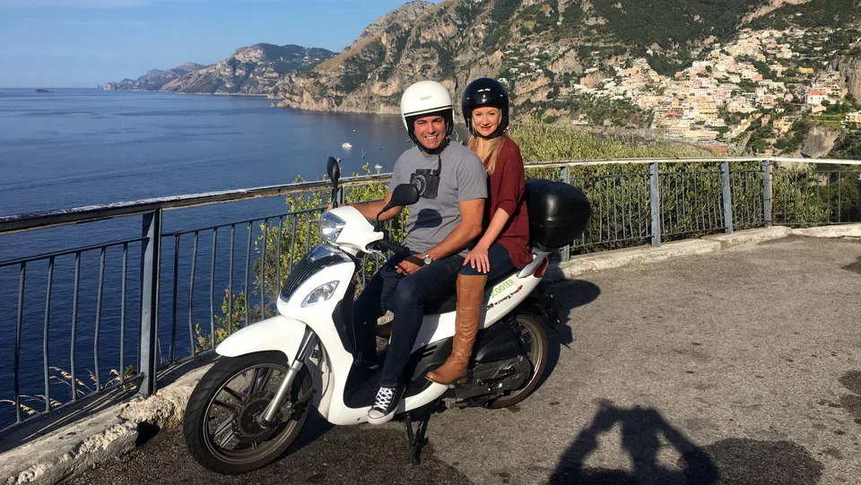 Alex and Kristin wearing helmets on a scooter with Positano and the coast behind them