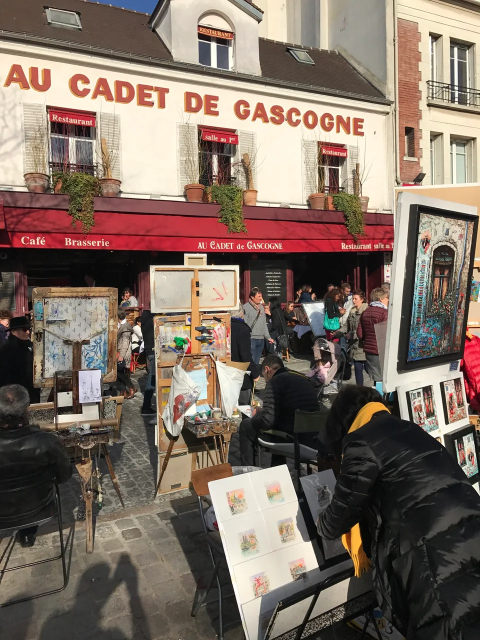 Os toldos vermelhos e a placa do Au Cadet de Gascogne junto a pinturas ao ar livre