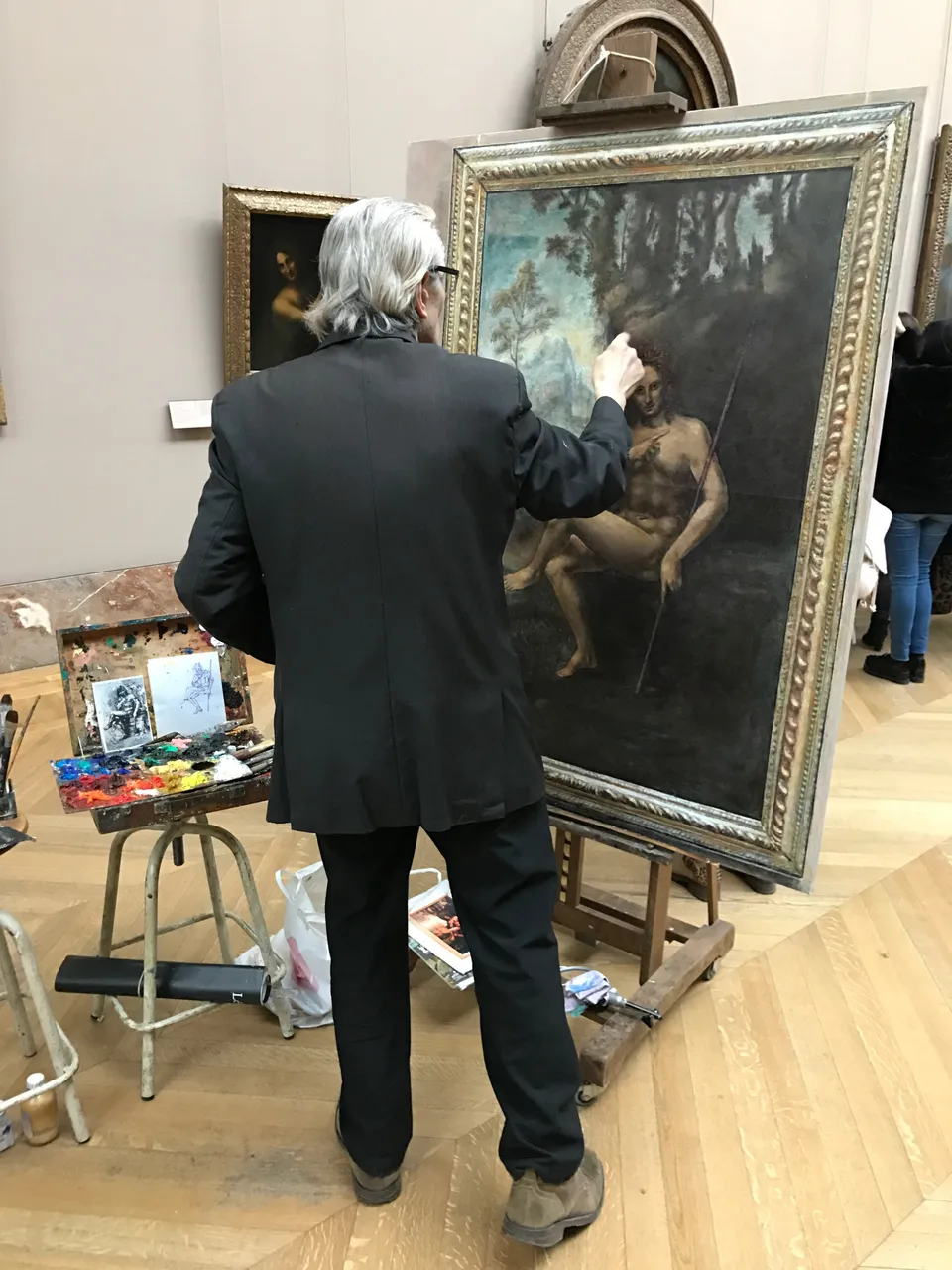 A painter working at an easel in front of a large painting inside the Louvre