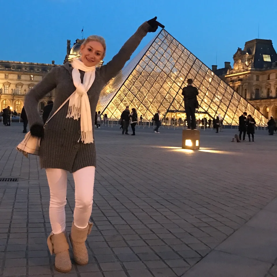 Kristin com as mãos levantadas perto da Pirâmide do Louvre iluminada ao entardecer