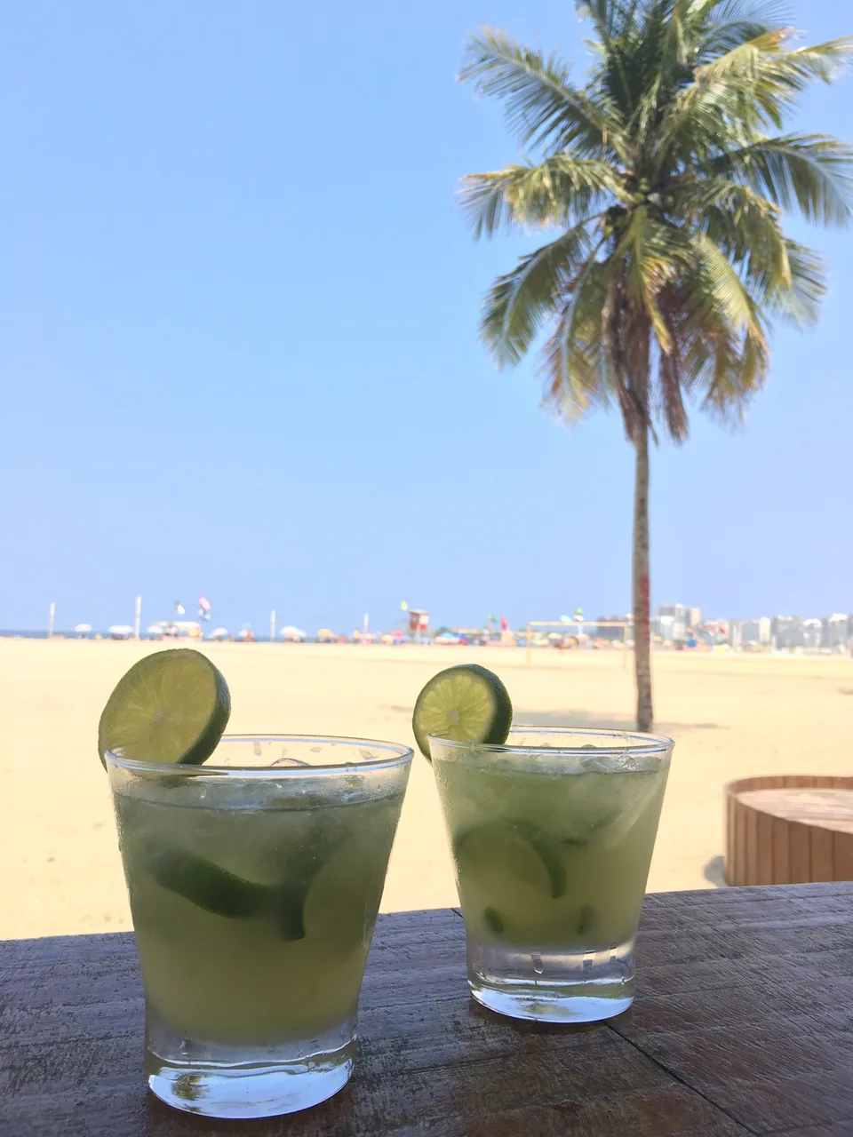 Two lime-garnished drinks on a beachside table with a palm tree beyond