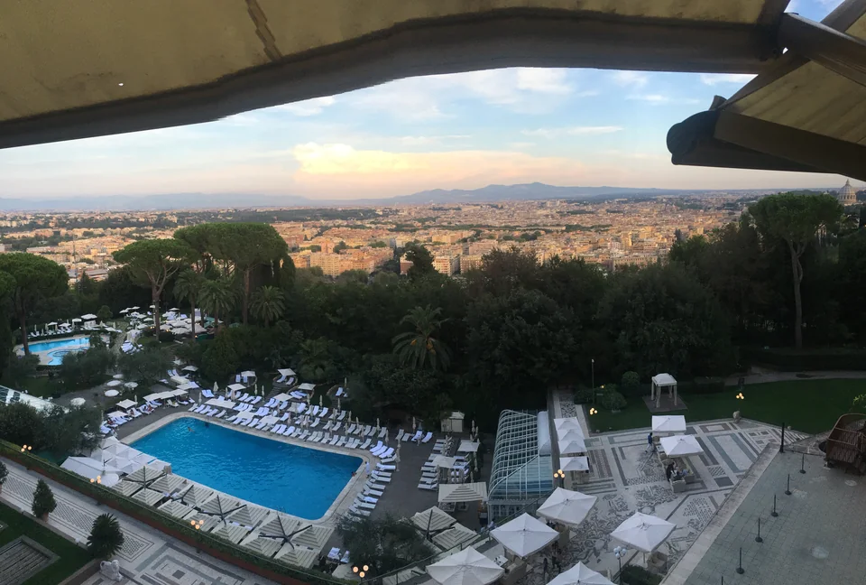 A wide view across Rome with a swimming pool and trees in the foreground