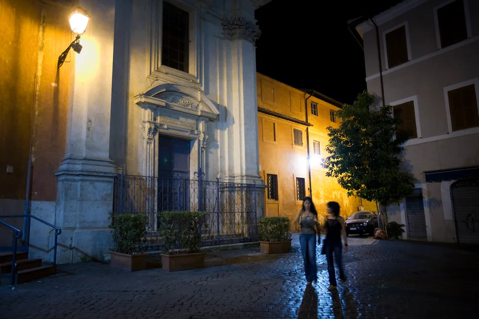 Duas pessoas atravessando paralelepípedos sob luzes quentes junto a uma igreja em Trastevere à noite